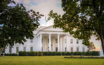 A large white house with columns and many windows. The house sits on a expansive green lawn with a large circular fountain and is framed by two green leafy trees. By Ana Lanza on Unsplash.