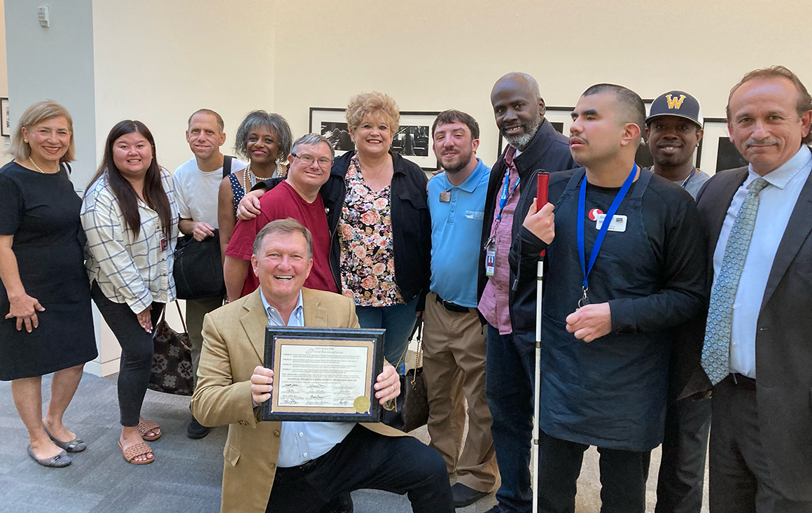 A group of people, including a person with Down syndrome and another with a visual impairment and walking cane, stand behind a kneeling man displaying a framed commendation.