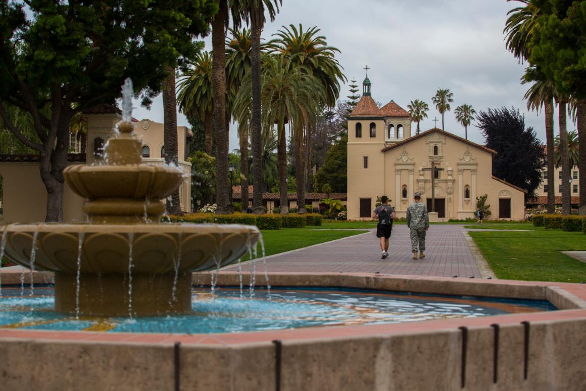 Fountain and gardens at a mission under a colorful sky.